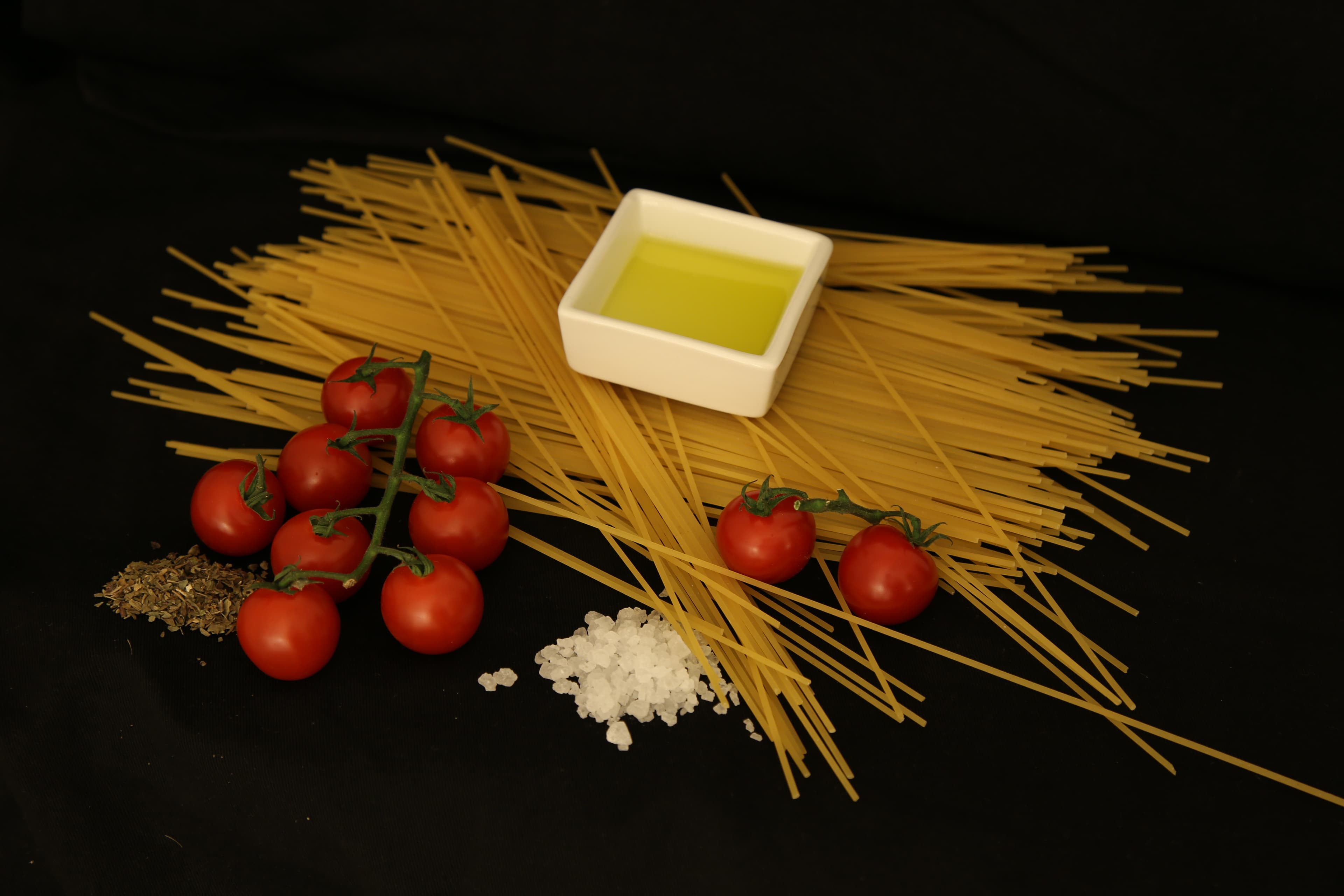 Pęk suchych spaghetti z pszenicy durum na czarnym tle, obok pomidorków, oliwy, soli i ziół